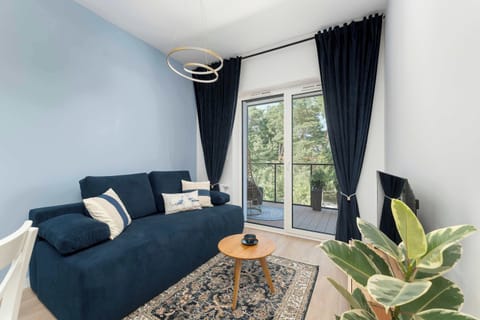 A bright living room features a blue sofa decorated with pillows. A low wooden table sits in front, while large windows reveal a tree-filled view. A potted plant stands in the corner on the wooden floor.