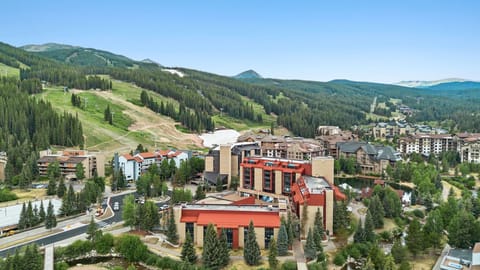 Aerial view of a mountain resort village nestled among forested slopes, with ski runs visible across the peaks and alpine community buildings dotting the landscape.