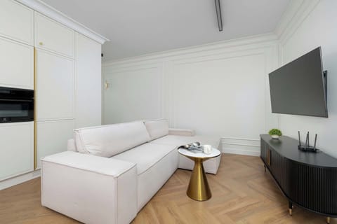 A modern living room featuring a white sofa, a black TV console, and a wall-mounted flat-screen TV. Sunlight filters through light curtains onto warm wooden herringbone flooring.