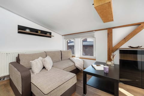 A cozy living room features a beige sectional sofa positioned around a dark coffee table. Exposed wooden ceiling beams add rustic warmth to the bright interior.