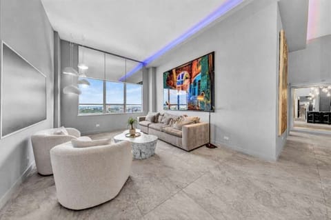 Modern loft-style living room with soaring ceilings, plush swivel chairs, and a cozy sofa arranged around a marble coffee table. Vibrant artwork, LED accent lighting, and skyline views through oversized windows create a chic, welcoming retreat.