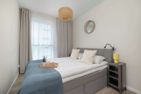 A cozy bedroom with a double bed made up with light linens and a blue throw. Bright daylight enters through a window draped with sheer and gray curtains.