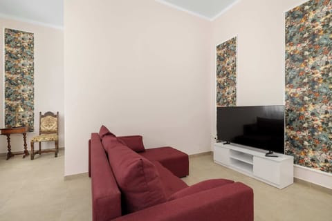A living area features a dark red corner sofa facing a flat-screen TV on a minimalist white cabinet. Floral patterned wallpaper strips frame the wall behind the set up, while smooth walls and light flooring create a bright environment.