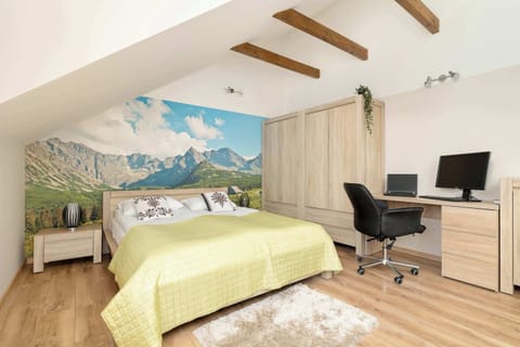 An attic bedroom features a double bed with a yellow spread in front of a scenic mountain wall mural. A study desk with an office chair sits in the corner.