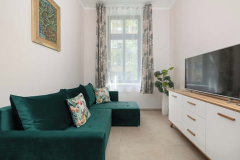 A living room with a green sofa and a TV on a white cabinet. Large windows make the space bright.