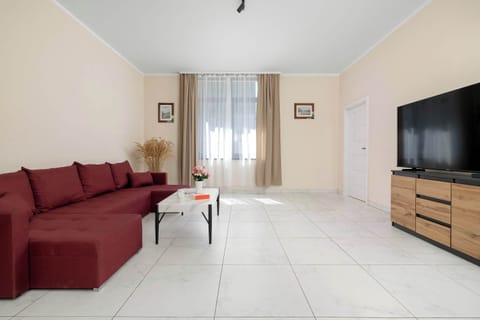 A bright living room with a red corner sofa, a coffee table, and a TV. Large windows fill the space with natural light.