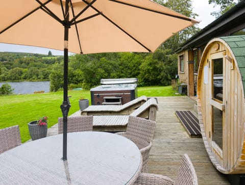 Decking that runs the length of the lodge, with a table under the parasol and the hot tub only a few steps away. The water sits just beyond the lawn.