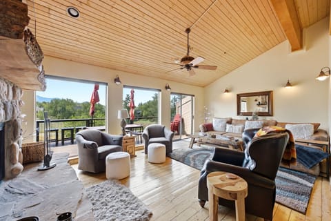 Bright, open concept living space featuring wood paneled ceilings and cozy fireside seating, opening directly onto a scenic outdoor deck