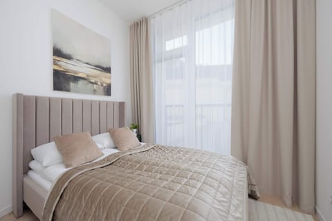 A cozy bedroom features a double bed with a tufted headboard and beige bedding. Sheer curtains hang beside a large window, filling the space with soft light. Above the bed hangs subtle modern artwork.