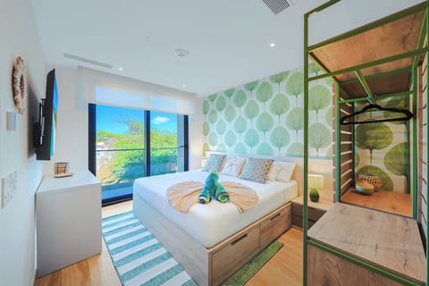 Unwind in your coastal-inspired bedroom with bright tropical wallpaper, a cozy bed adorned with playful towel art, and lush garden views framing your private retreat.