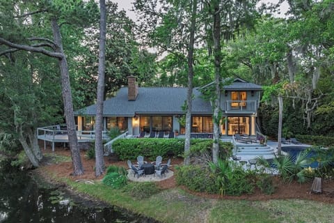 67 Plantation Drive, nestled among towering trees, featuring expansive windows, multiple outdoor decks, pool, and a cozy firepit seating area—an inviting woodland retreat.