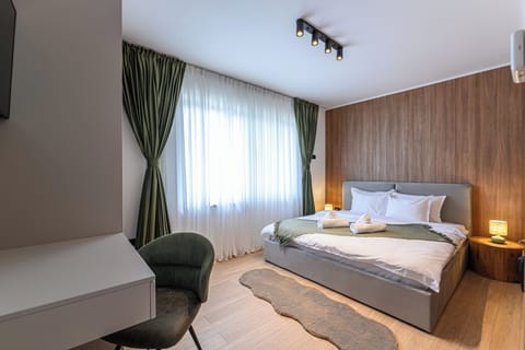 Queen-size bedroom with upholstered grey headboard, warm oak slatted wall panel, dark accent chair, room-darkening green curtains and sheer layer for natural light. Grand Chalet Suite 2, Poiana Brasov.