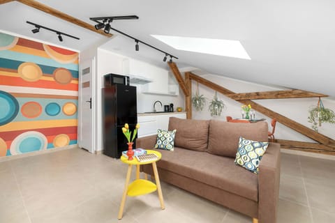 A view of the living area highlights a brown sofa, a small yellow side table, and vibrant, multi-colored circular pattern wallpaper. Exposed wooden ceiling beams run across the top of the room.