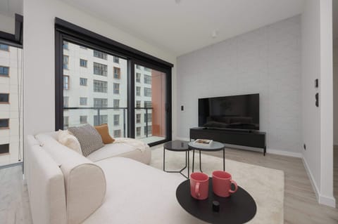 A bright living room features a comfortable white corner sofa and a black TV stand. Large windows let in plenty of natural light, illuminating the light wooden floor.