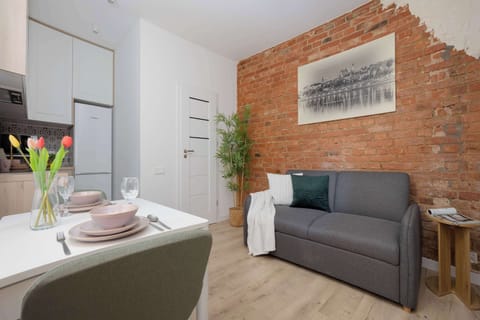 A gray sofa stands against an exposed brick wall with framed artwork above it. A dining table and kitchenette are visible nearby.