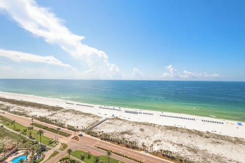 Aerial view captures the pristine Gulf Coast beachfront with white sand shores, turquoise waters, and palm-lined coastal pathway stretching along the shoreline.