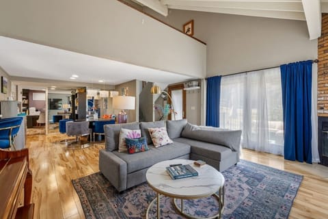 Bright and spacious open-plan living room with vaulted ceilings, comfortable sectional seating, hardwood floors, and abundant natural light flowing into the adjoining dining and kitchen spaces.