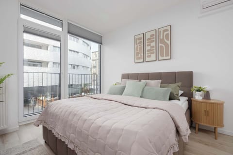 A cozy bedroom features a double bed dressed in light pink and sage green linens beneath triptych wall art. Large windows look out onto surrounding buildings.