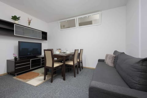 A living room with a sofa, dining table, and TV creates a comfortable shared space. Large windows make the room bright and inviting.