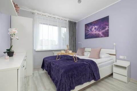 A bright and cozy bedroom with a comfortable double bed and soft purple accents. Large windows fill the room with natural light, creating a relaxing atmosphere.