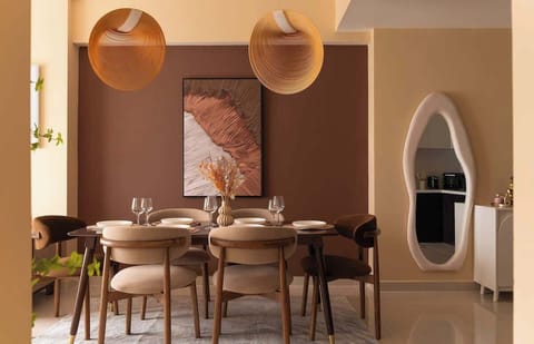 Striking dining space with bold chocolate accent wall, pendant lights, and sculptural centerpiece. A mix of soft velvet chairs surrounds a table set for 6. The arched mirror and kitchen peek complete the design-forward, Instagrammable vibe.
