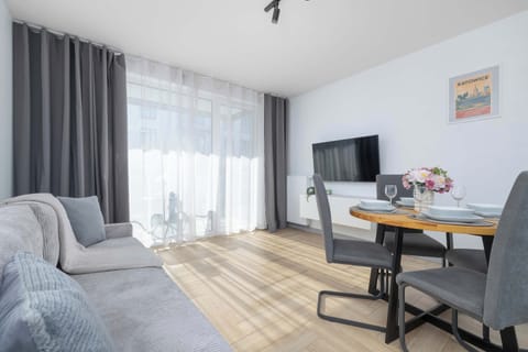 A bright living room features a comfortable grey sofa and a small dining table set for two. Large windows let in plenty of natural light.
