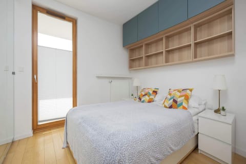 A compact bedroom with a double bed, built-in wooden shelving above the headboard, and a large window bringing in natural light.