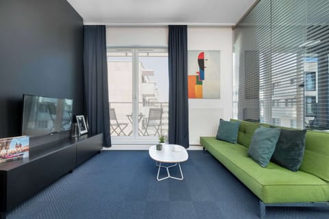 A modern living room featuring a bright green sofa, a small coffee table, and a television. Natural light streams in through the glass balcony doors.