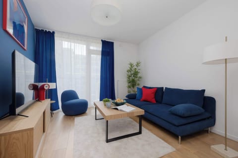 A bright bedroom with a comfortable blue sofa, coffee table, and colorful accents. Large windows fill the space with natural light, creating a welcoming atmosphere.