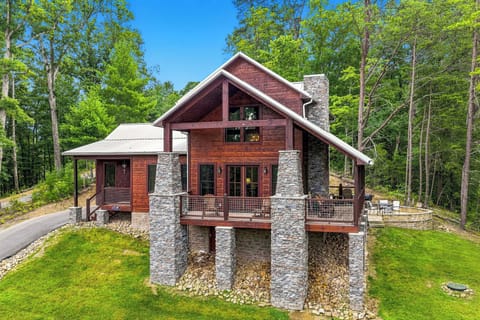 A stunning vacation home with rustic wood siding and stone accents nestled in a lush forest. The property features expansive decks and charming architectural details, surrounded by vibrant greenery and clear blue skies.
