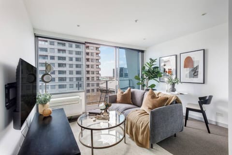 Relax in the bright, modern living area with a soft grey sofa, glass coffee table, and decorative greenery, all set against sweeping views of West Melbourne’s rooftops and leafy treetops.