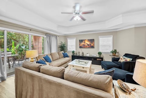 Unwind in this sunlit living space featuring a soft beige sectional and a cozy blue armchair. The stylish decor and modern fireplace create a warm atmosphere, complemented by greenery visible through the glass doors.