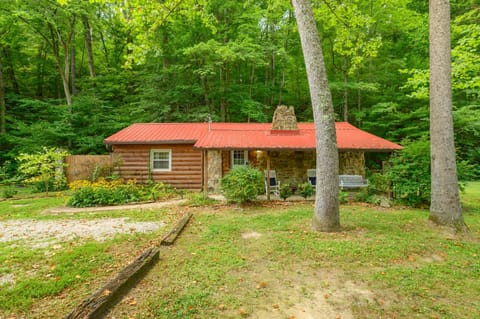 Front view of a rustic log cabin with a red metal roof, stone chimney, covered porch with rocking chairs, and lush green trees.