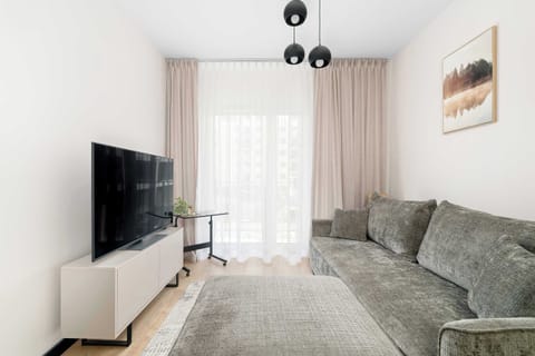 A bright living area featuring a large patterned sofa, a wall-mounted television on a minimalist TV stand, and long neutral curtains framing a sunny window. Light wood flooring enhances the open feel of the space.