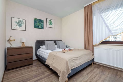 A bright bedroom with a comfortable double bed, a wooden dresser, and decorative artwork on the wall. Large windows with curtains bring plenty of natural light into the room.