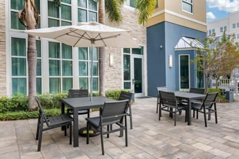 Outdoor patio with metal tables, chairs, and white umbrellas beside a palm-lined wall