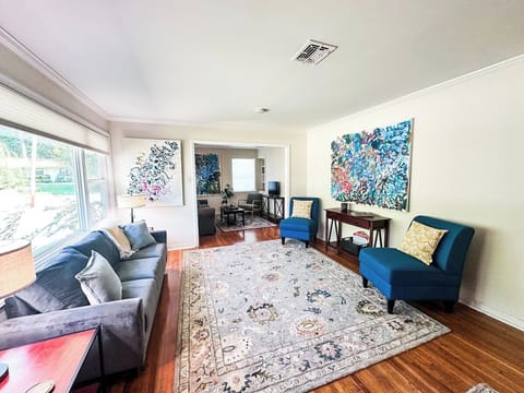 Unwind in this bright and spacious living room, featuring a plush grey sofa, vibrant teal accent chairs, a large patterned area rug, colorful abstract wall art, and an open entryway leading into an adjoining sitting area.