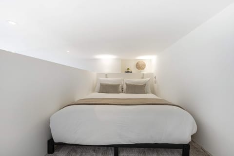 Rest in the minimalist lofted sleeping alcove featuring a double bed dressed with crisp white bedding, tan throw blanket, and neutral accent pillows.