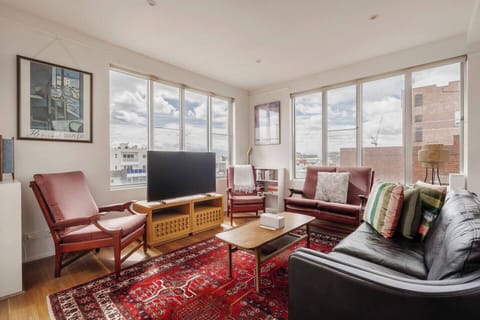 Unwind in a sunlit lounge framed by expansive windows, with mid-century seating, a leather sofa, air conditioning, heating, a TV, and shelves of books for quiet evenings.