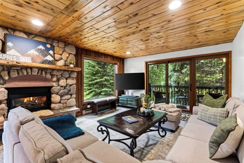 Cozy up by the stone fireplace in this inviting living room, framed by warm wood details and peaceful forest views.