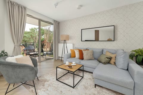 A bright living room features a light grey corner sofa styled with mustard throw pillows, a glass coffee table, and an armchair. Sliding glass doors lead onto a balcony.