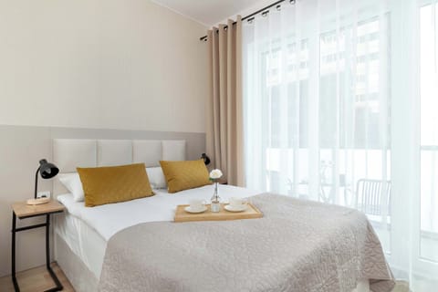 A cozy bedroom area with a double bed dressed in white bedding and accented with mustard-yellow cushions, positioned beside a large window.
