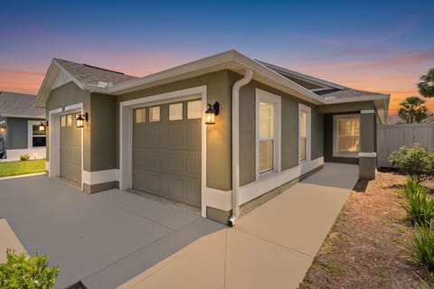 The property greets you at dusk with warm exterior lighting and neatly landscaped surroundings, set against a stunning pastel sky and swaying palms.