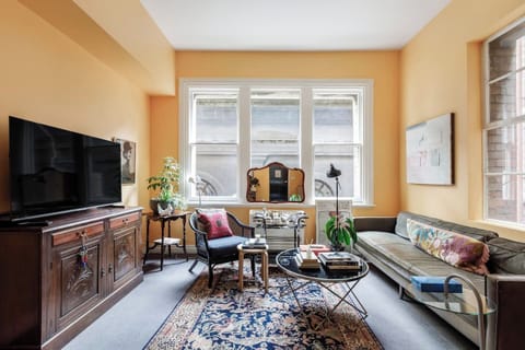 Relax into the mid-century styled living area featuring plush seating, curated books, lush plants, a TV, and large sash windows that fill the space with light.