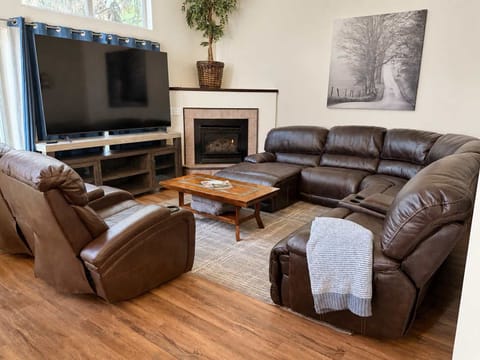 Open-concept living and dining space with seating for 6, leather recliners, a large TV, fireplace, and tall windows that fill the room with natural light.