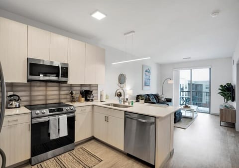Modern kitchen with light wood cabinetry, stainless steel appliances, and a spacious island overlooking the living area. Bright and open layout flows to a cozy seating area and private balcony with city views.