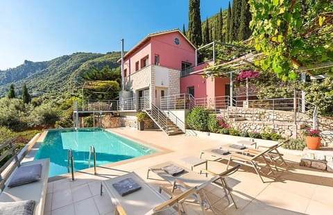 An elevated view showcases a pink hillside villa featuring an outdoor swimming pool, patio lounge chairs, and lush greenery with a mountain backdrop.