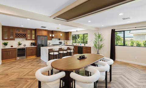 Dining area features an oval table surrounded by white chairs, positioned in front of a the kitchen equipped with a breakfast island, appliances and utensils. 