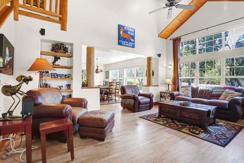 A spacious, rustic-style living room features warm leather seating, light hardwood floors, and towering vaulted ceilings with large windows that look out into a lush wooded landscape.