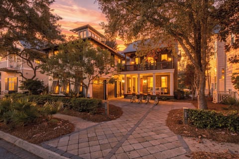 The property exterior welcomes guests at golden hour, with warm lights glowing through windows and mature trees framing the entrance—a peaceful coastal neighborhood setting to begin your stay.